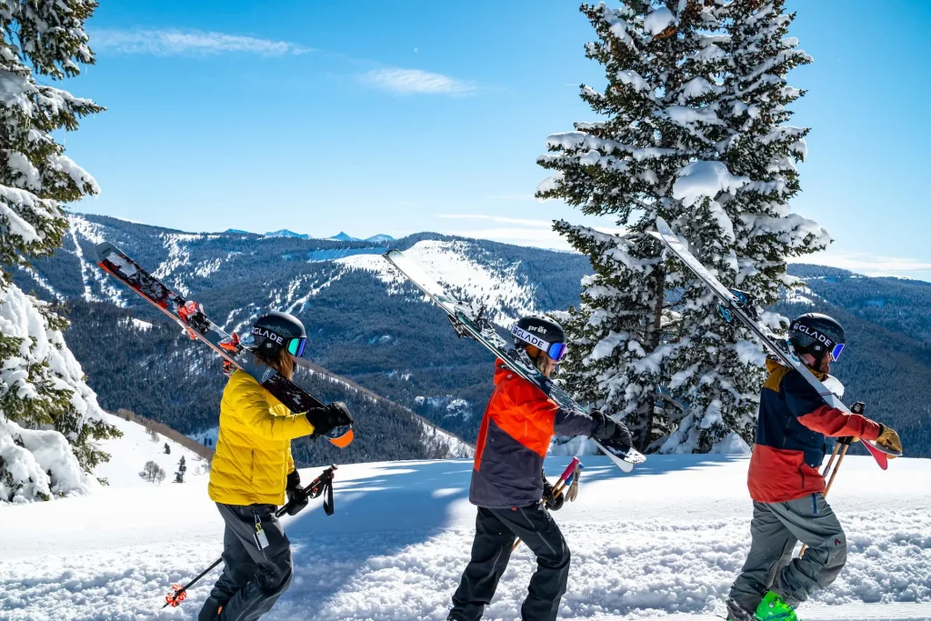 Three skiers in colorful winter gear walking through a snowy mountain landscape with snow-covered trees and distant peaks.