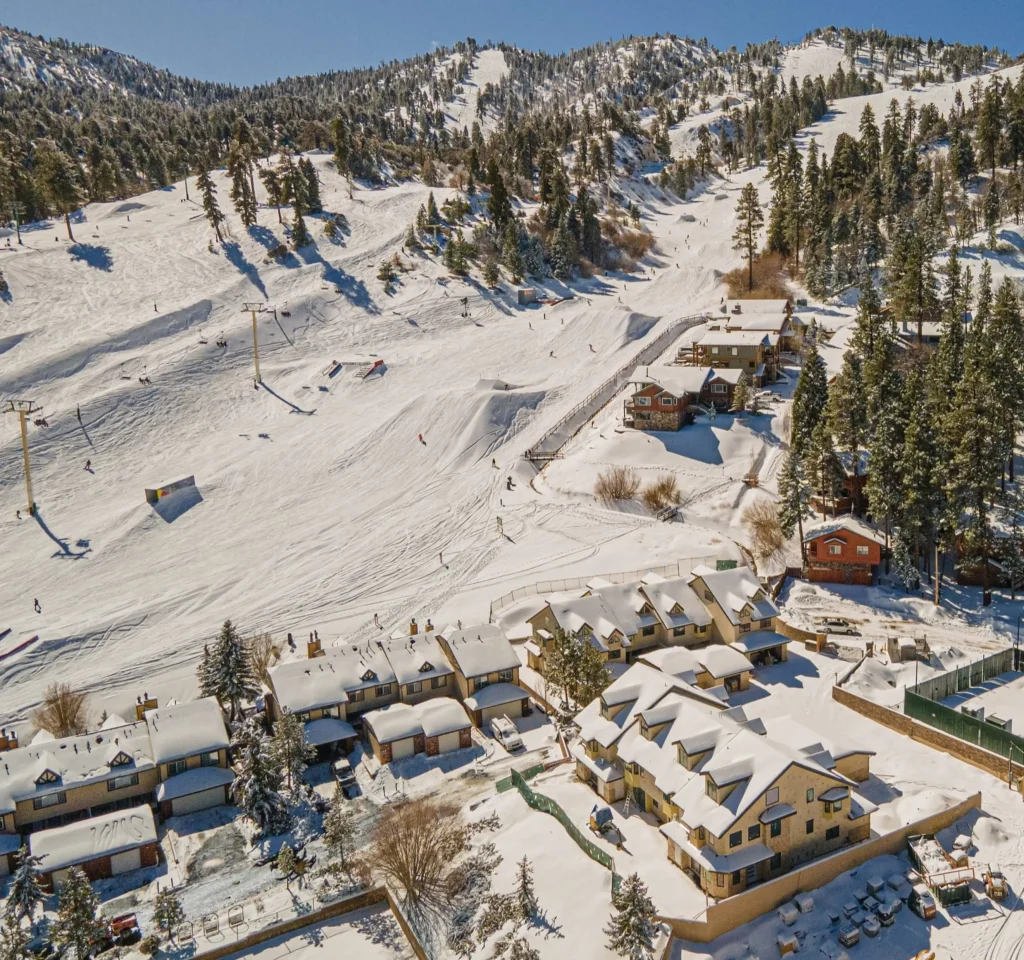 Drone view of snow-covered cabins and ski slopes surrounded by pine trees and mountains under a clear blue sky at Big Bear Mountain.