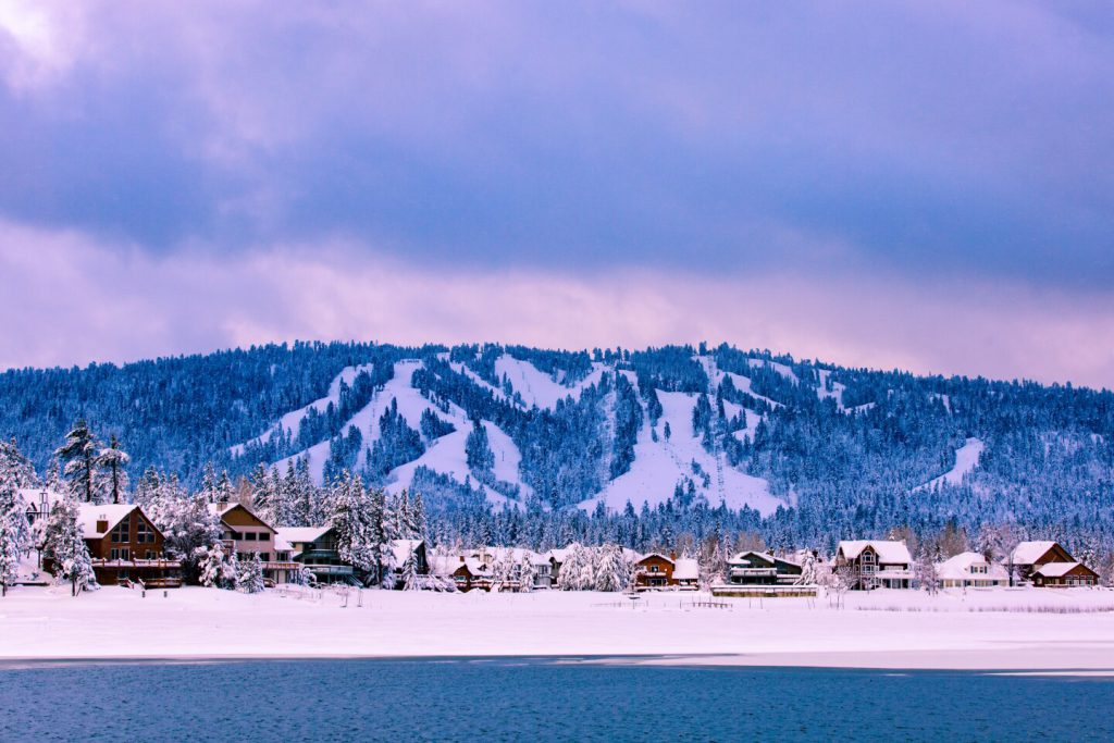 Snow-covered mountain with ski trails, pine forest, lakeside village, and a partly cloudy sky with soft hues.