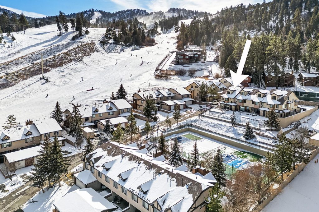 Aerial view of snow-covered townhomes beside Bear Mountain Ski Resort with skiers on the slopes and a white arrow highlighting a specific property.