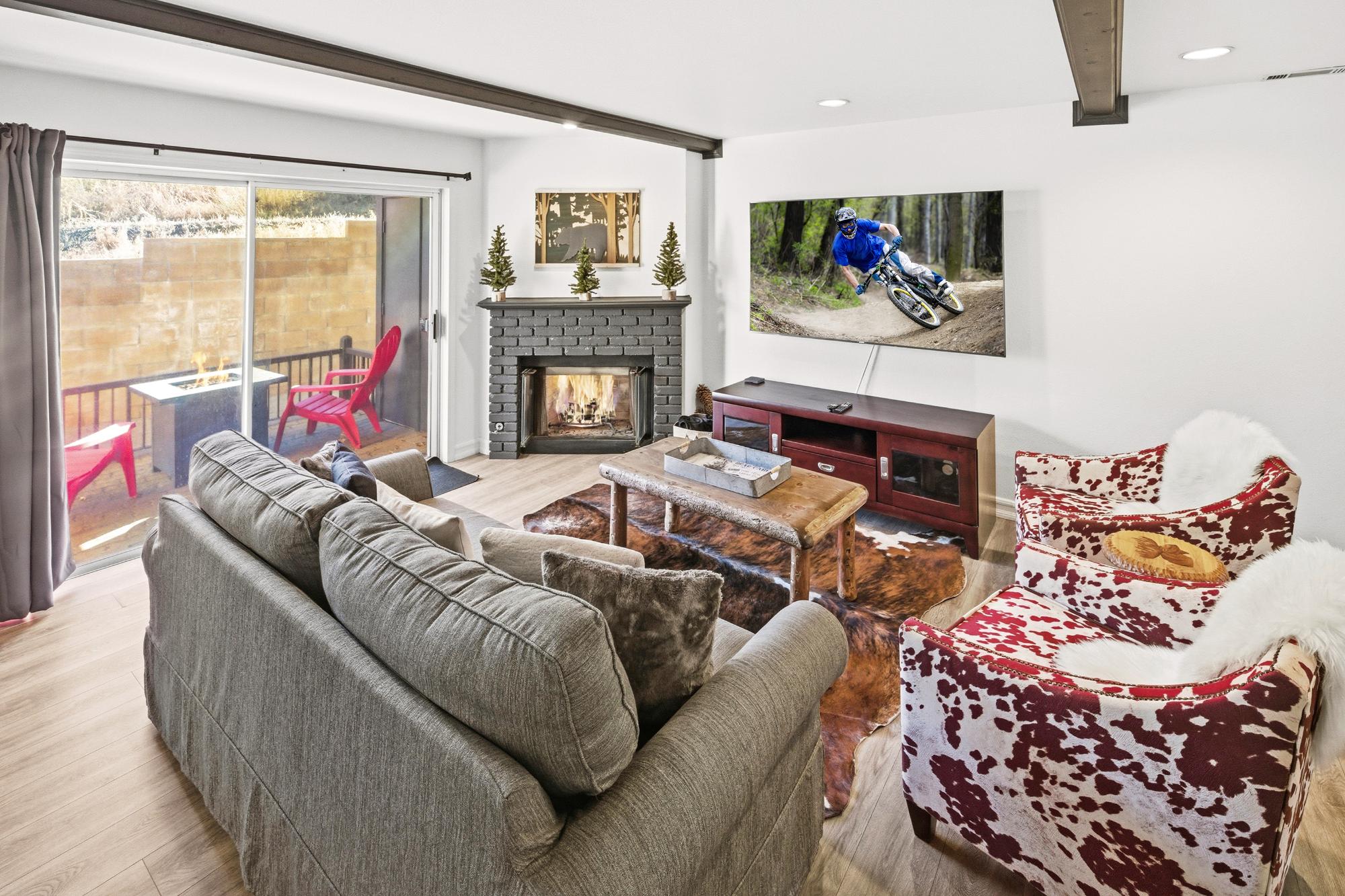Modern living room with a fireplace, TV displaying a mountain biker, and cozy seating, including a cowhide armchair. Large windows offer a view of outdoor seating, and the room features wooden flooring.