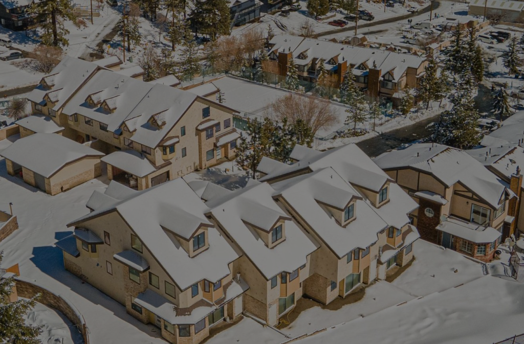 Aerial view of snow-covered townhomes near Bear Mountain Ski Resort surrounded by pine trees and mountain scenery on a sunny winter day.