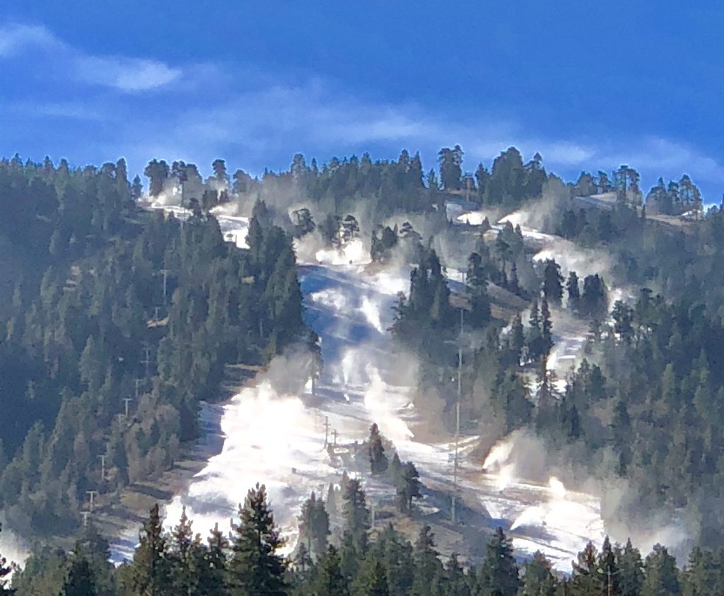 Snow machines cover the Bear Mountain ski slopes with fresh powder, surrounded by pine trees and a clear blue sky on a bright winter morning.