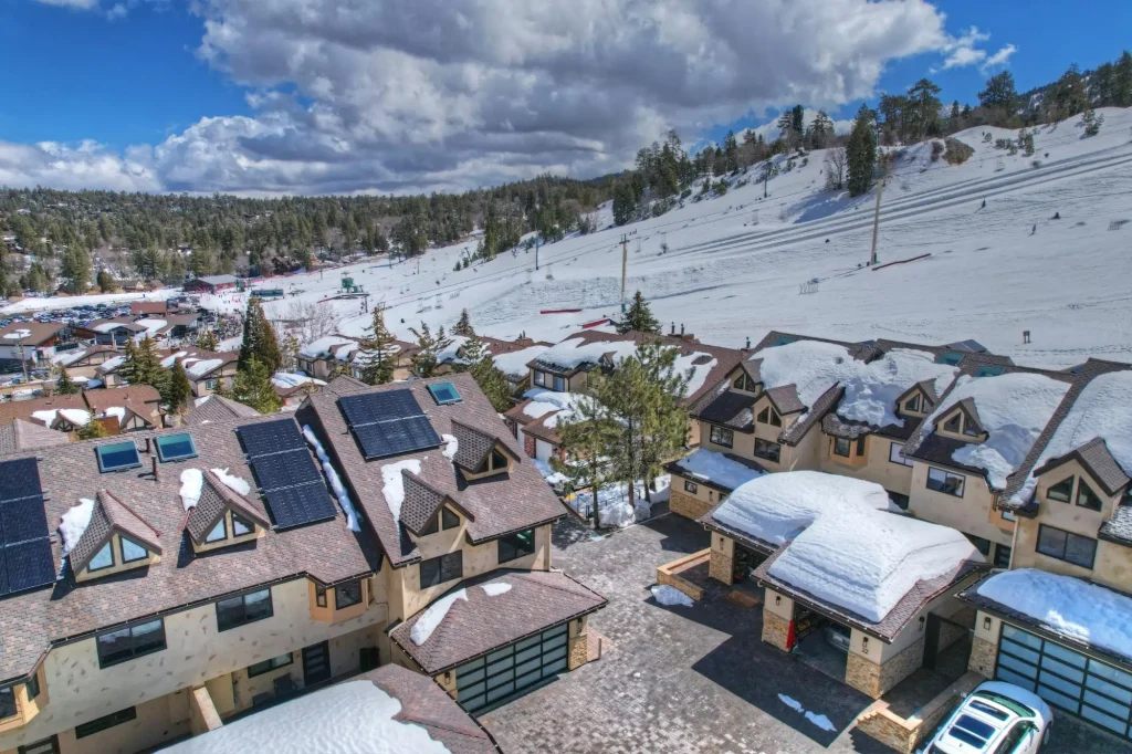 Aerial view of snow-covered condos with solar panels located beside ski slopes and surrounded by pine trees at Bear Mountain in Big Bear, California.