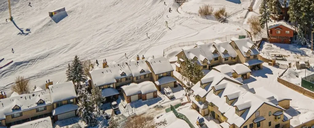 Aerial view of snow-covered vacation condos situated directly at the base of Bear Mountain Ski Resort in Big Bear, California, with skiers visible on the slopes nearby.