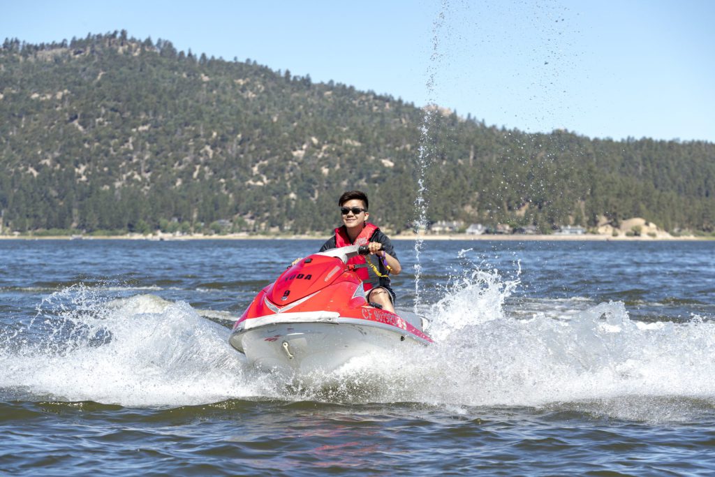 Person riding a red and white Yamaha jet ski on a lake with forested mountains and shoreline buildings in the background under a sunny sky.