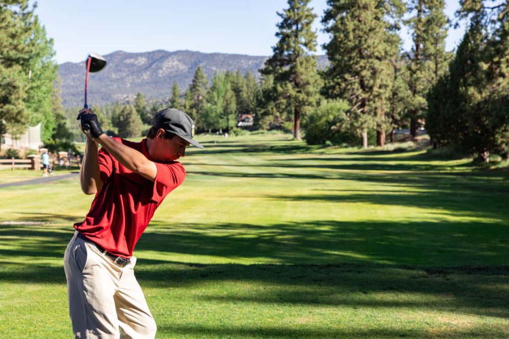 A golfing in a red shirt and khaki pants with a black golfing glove is in mid swing looking down at his golf ball as he is about to drive the ball down the golf course surrounded by pine trees and mountains.