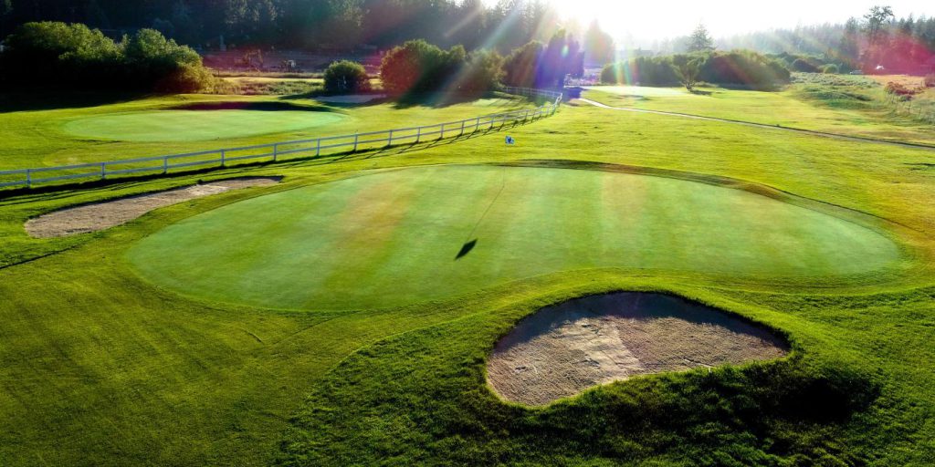 Very green golf course hole with two surrounding sand traps and another hole in the upper right. The sun is beaming down on the course.