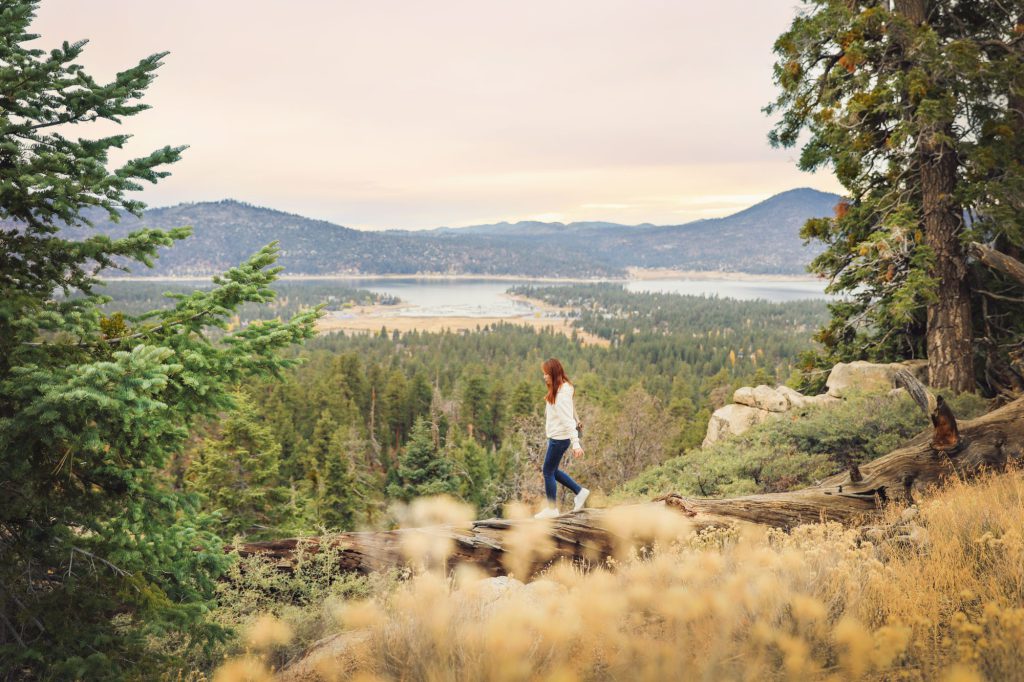 A woman hiking on a forested trail with a scenic view of a lake and mountains in the distance.