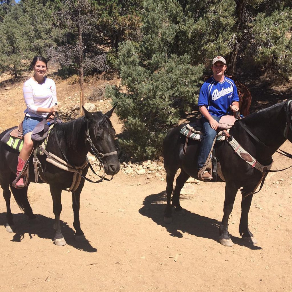 A man and woman riding horses along a dirt trail surrounded by trees, both smiling and enjoying the horseback riding experience.