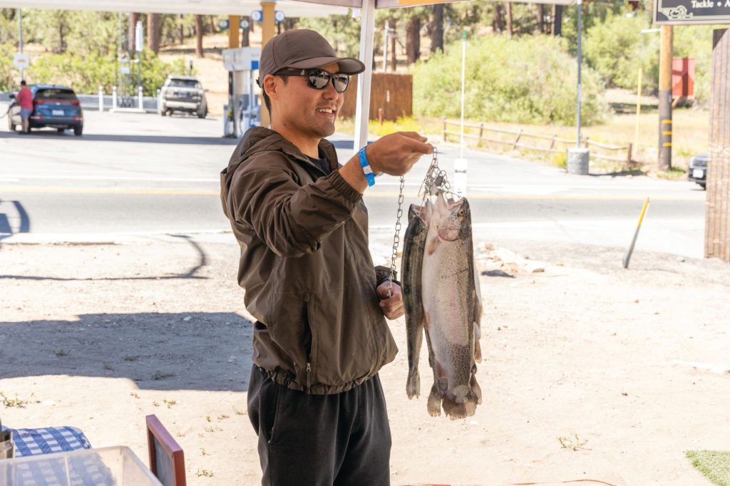 A man holding two freshly caught trout by their tails with a proud smile, standing under a shaded tent at a fishing spot.