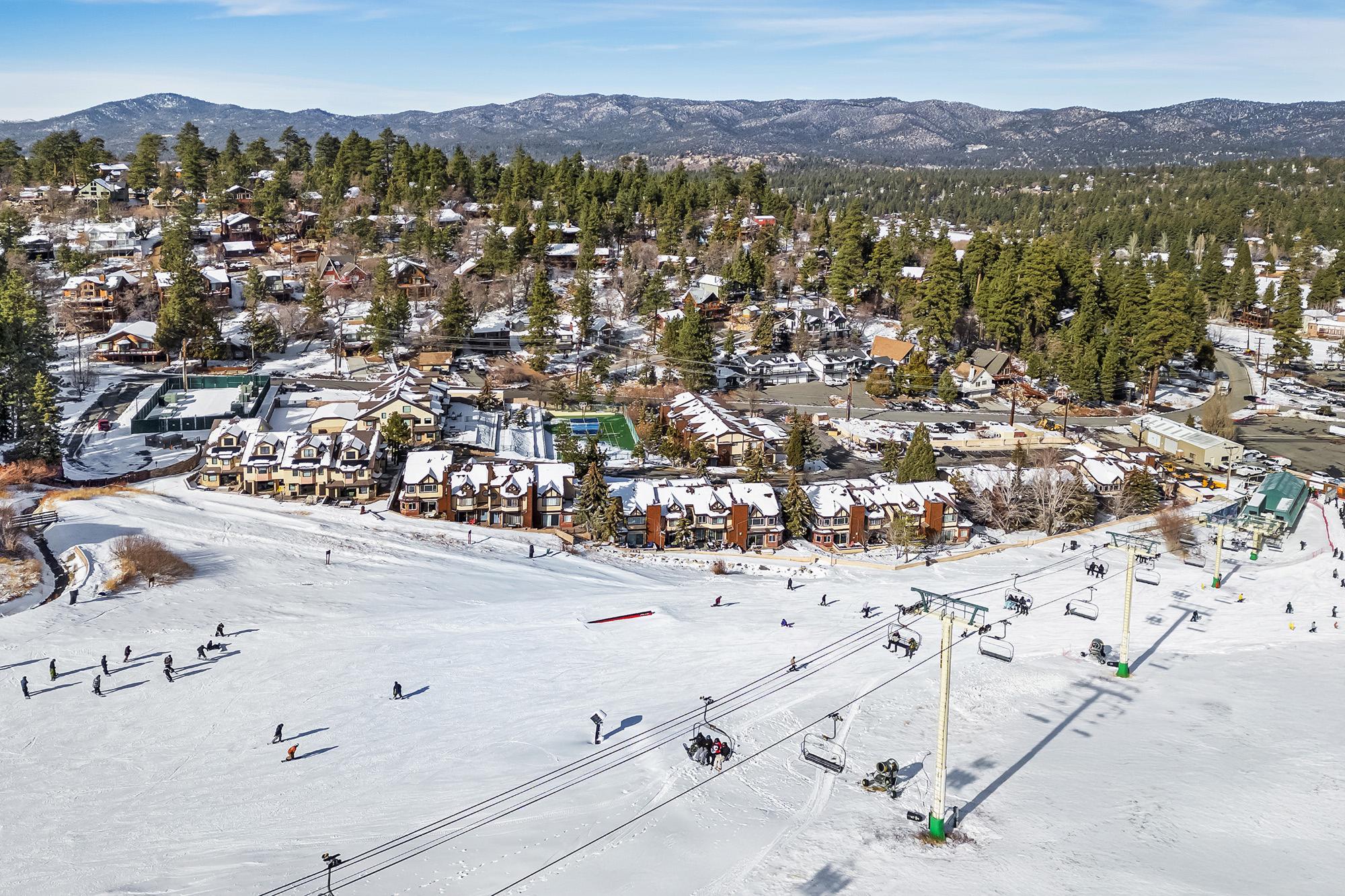 Aerial view of a snowy ski resort with chairlifts, skiers on the slopes, and mountain cabins surrounded by pine trees