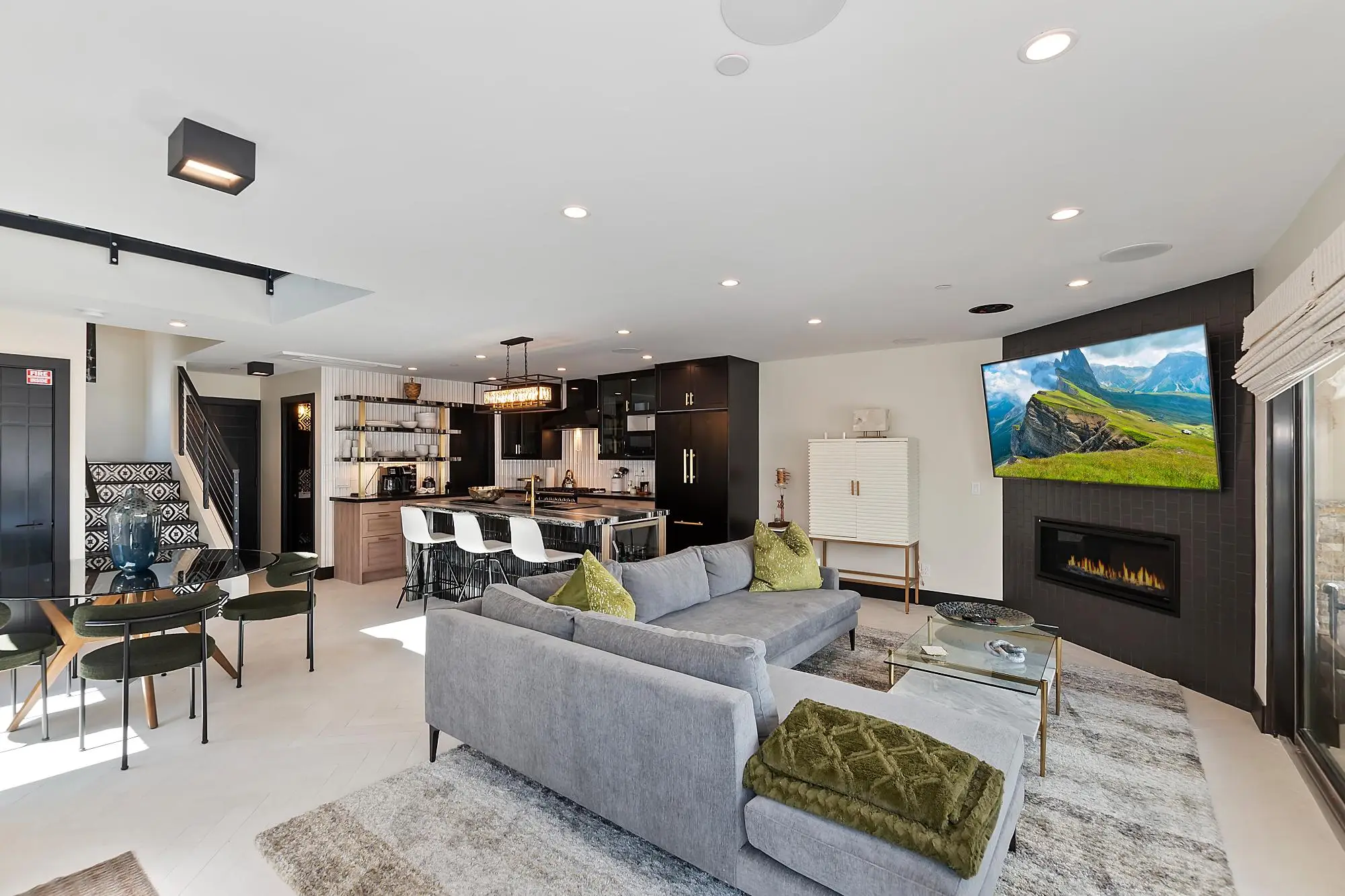 Open-concept living space with gray sectional, green ottoman, fireplace, and kitchen-dining area in background.