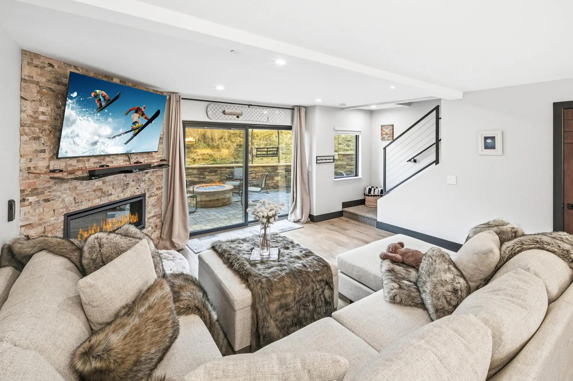 Modern mountain-style living room with stone fireplace, wall-mounted TV, cozy fur throws, and sliding glass doors opening to a patio with fire pit.