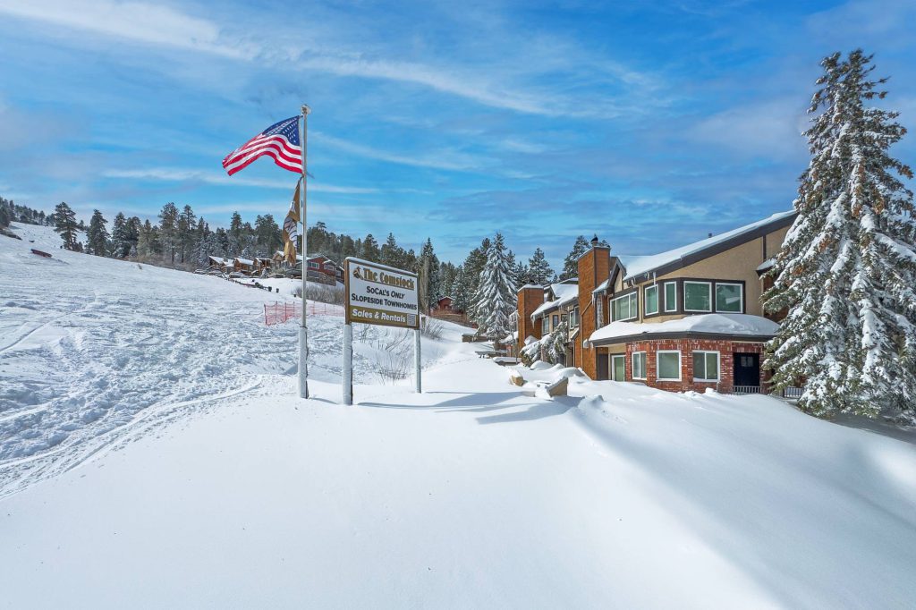 Snow-covered slopeside townhomes with an American flag and a sign reading “The Comstock” beside a ski run under a blue sky.