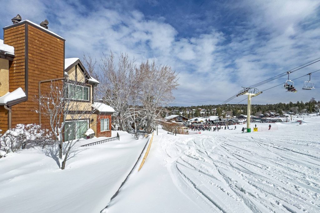 Ski lift and skiers passing directly beside snow-covered townhomes at a slopeside resort.