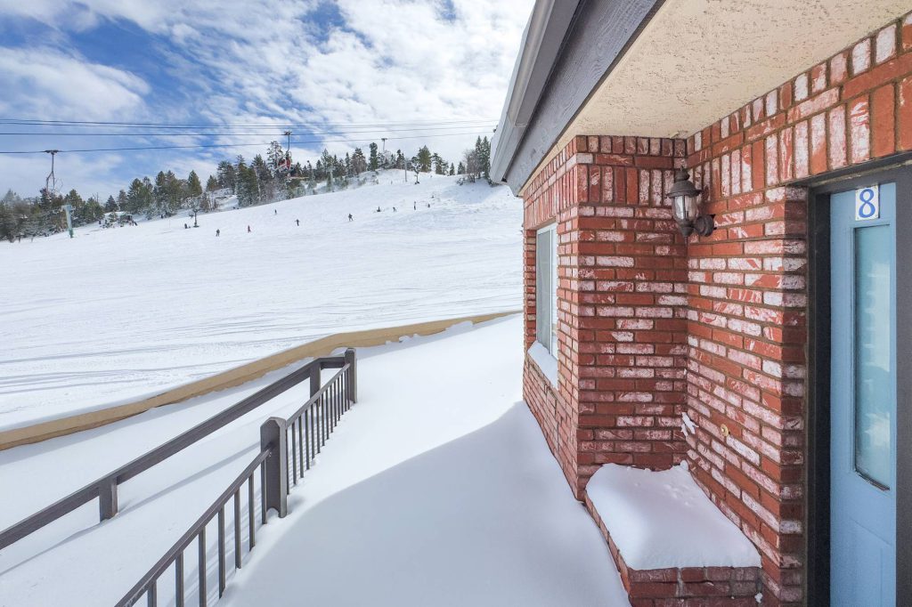 Balcony and brick exterior of a slopeside townhome overlooking a snowy ski run with skiers and a chairlift.