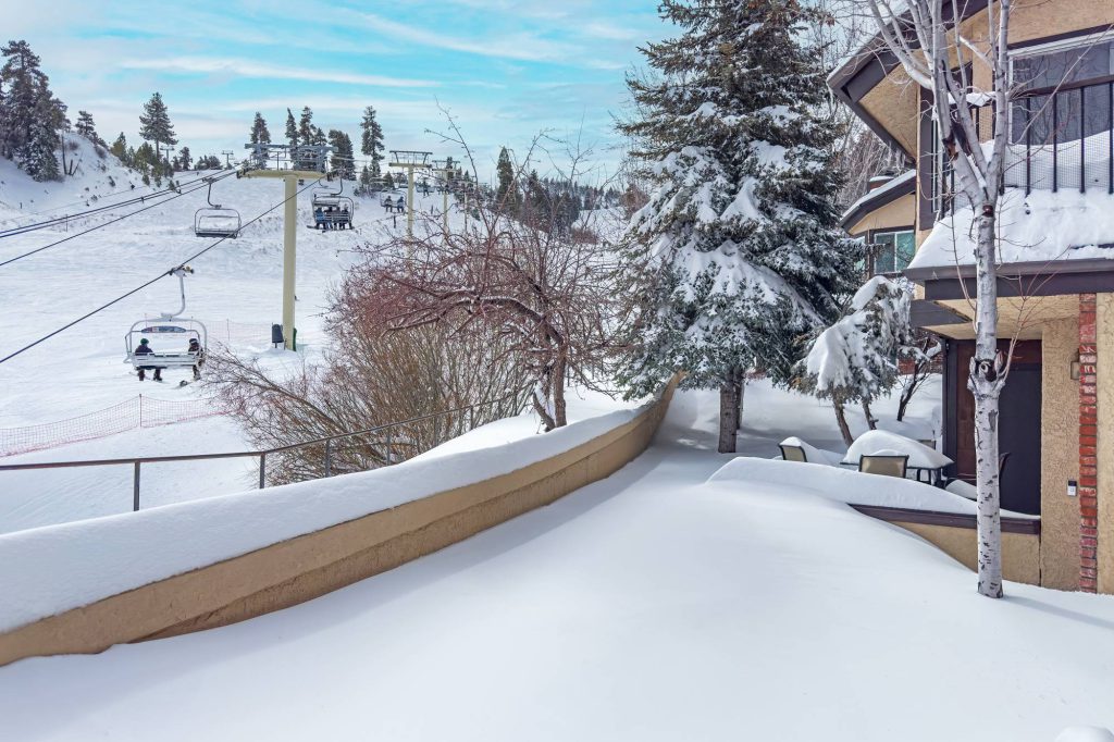 Snow-covered patio and retaining wall beside slopeside townhomes with a chairlift and ski slope in the background.