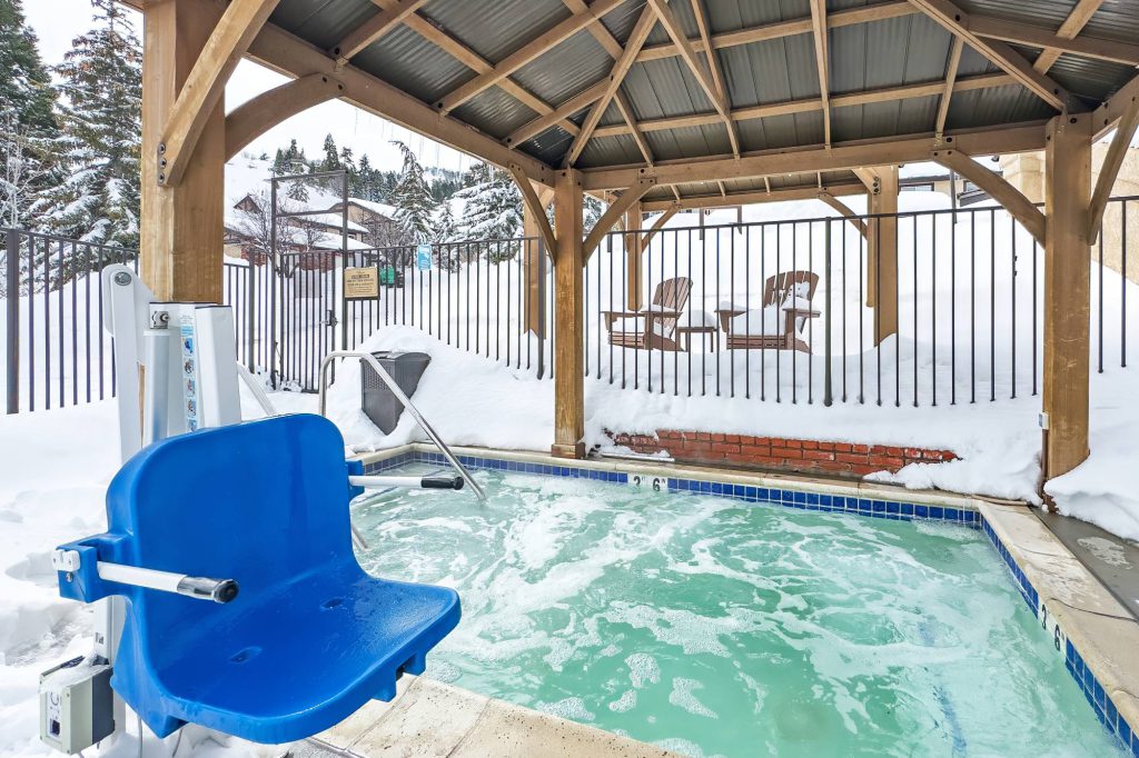 Outdoor heated pool and hot tub area with accessibility lift, surrounded by snow and covered seating.