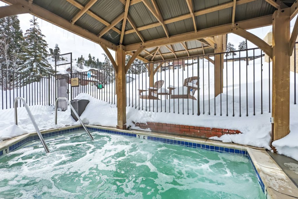 Covered outdoor hot tub with bubbling water, snow piled around the deck, and lounge chairs behind a black metal fence.