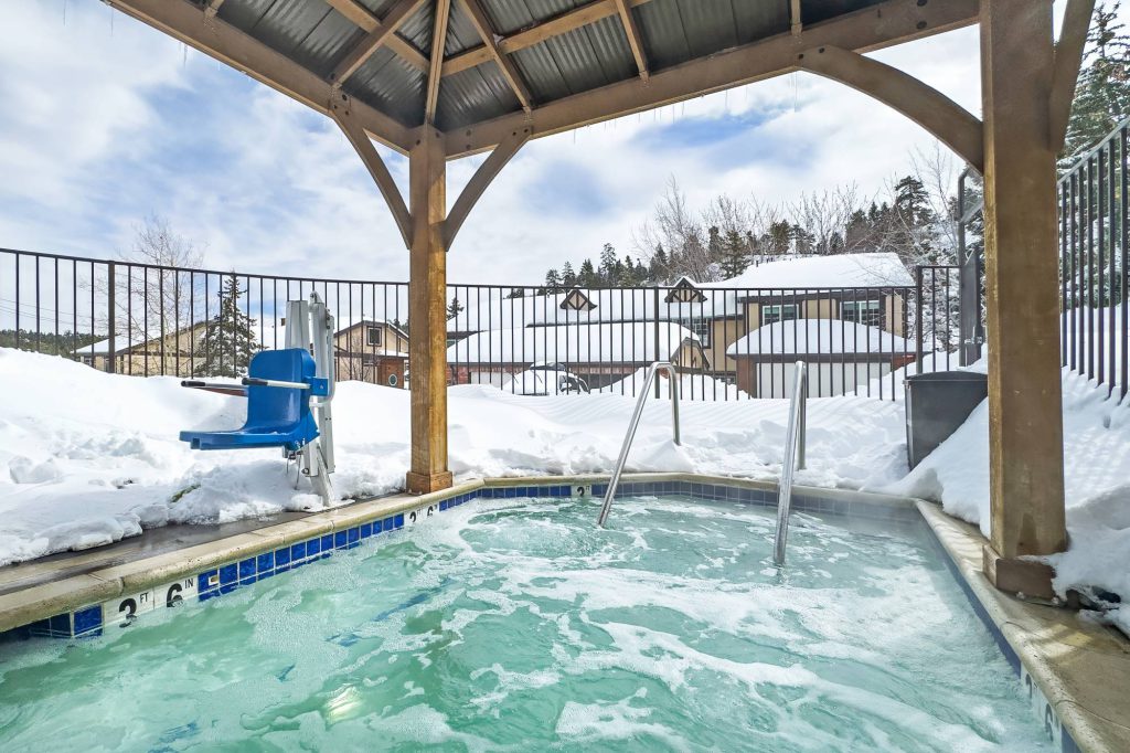 Covered outdoor hot tub with handrails and an ADA pool lift, surrounded by snow and slopeside buildings in the background.