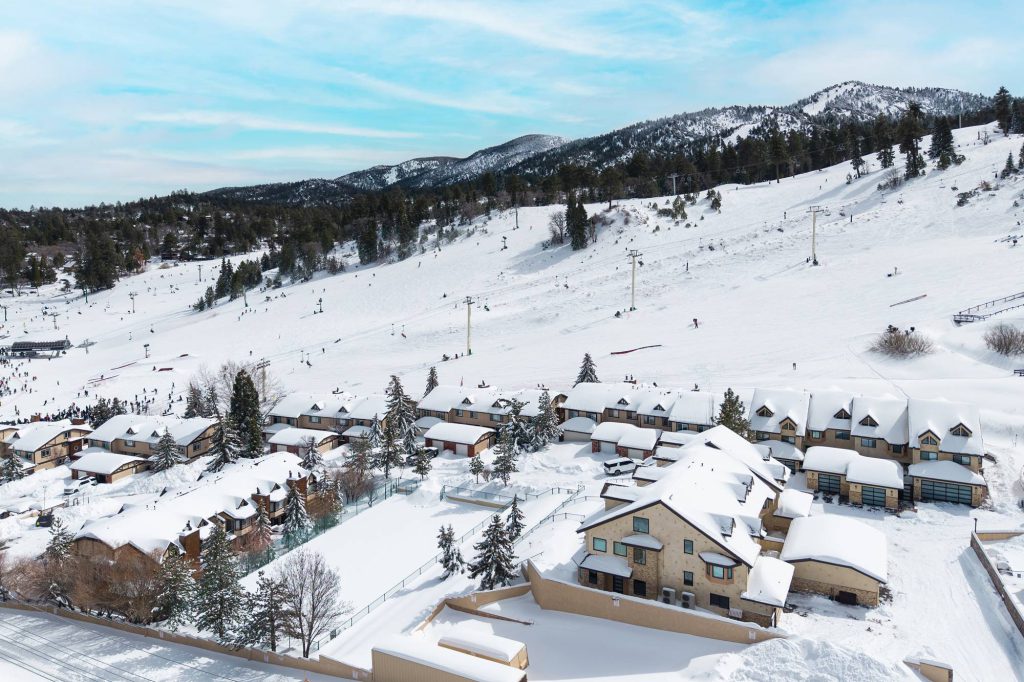 Aerial view of snow-covered slopeside townhomes lined up below a ski slope with chairlifts and skiers on the mountain.