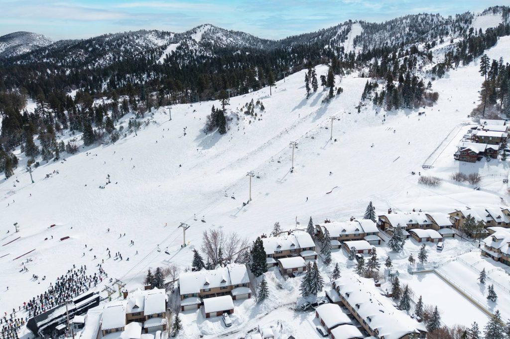 High aerial view of a ski resort base area with chairlifts, skiers, and snow-covered townhomes along the lower slopes.
