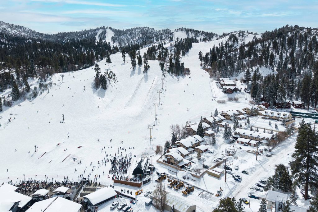 Aerial view of a ski lift line rising up the mountain with skiers below and snow-covered townhomes clustered at the base.