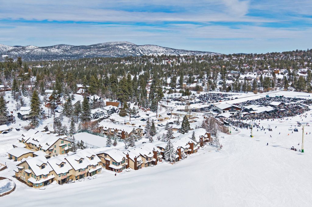 Aerial view of The Comstock slopeside townhomes beside a busy ski resort base area with chairlifts, skiers, parking lots, and snow-covered mountains in the background.