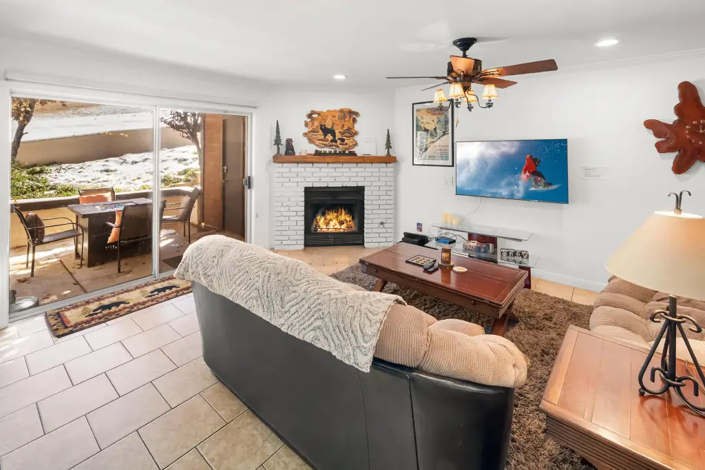 Cozy living room with white brick fireplace, wall-mounted TV, ceiling fan, and sliding glass doors leading to a snow-covered patio.